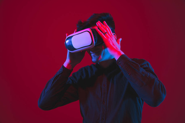 Caucasian mans portrait isolated on red studio background in purple-blue neon light, emotional and beautiful, playing wore VR-headset - Foto, afbeelding