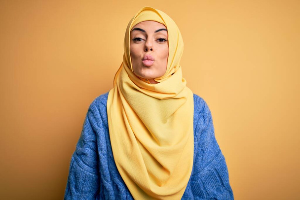 Genç, güzel, esmer, Müslüman kadın izole edilmiş sarı arka plan üzerinde Arap başörtüsü takıyor. Havada öpücük sallayan kameraya bakıyor. Güzel ve seksi. Aşk ifadesi. - Fotoğraf, Görsel