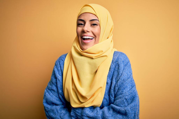 Fiatal gyönyörű barna muszlim nő visel arab hidzsáb felett elszigetelt sárga háttér mosolyog és nevetni keményen ki, mert vicces őrült vicc a kezét a test. - Fotó, kép