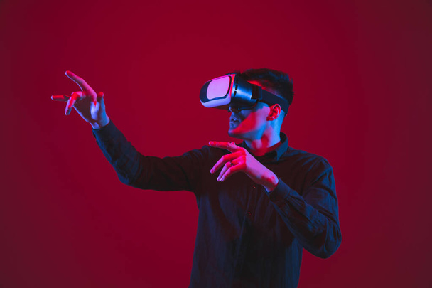 Caucasian mans portrait isolated on red studio background in purple-blue neon light, emotional and beautiful, playing wore VR-headset - Fotoğraf, Görsel