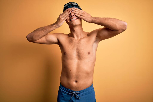 Young handsome man shirtless wearing swimsuit and swim cap over isolated yellow background covering eyes with hands smiling cheerful and funny. Blind concept. - Zdjęcie, obraz