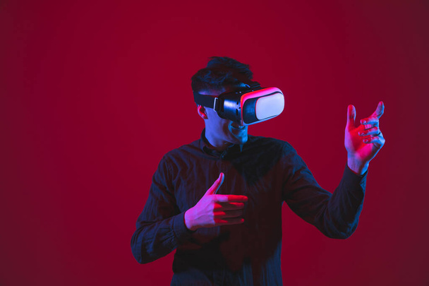 Caucasian mans portrait isolated on red studio background in purple-blue neon light, emotional and beautiful, playing wore VR-headset - Φωτογραφία, εικόνα