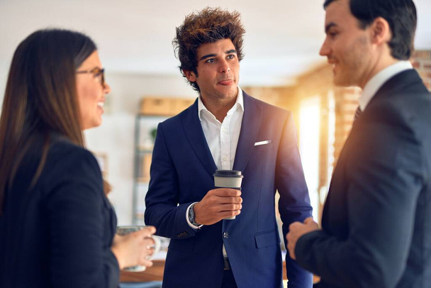 Молодая красивая группа бизнесменов улыбается счастливой и уверенной в себе, стоит с улыбкой на лице, пьет кофе и выступает в офисе
 - Фото, изображение