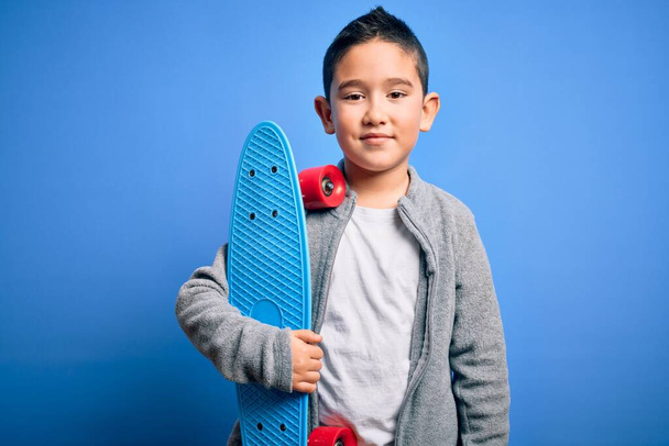 Young little boy kid skateboarder holding modern skateboard over blue isolated background with a happy face standing and smiling with a confident smile showing teeth - Фото, изображение