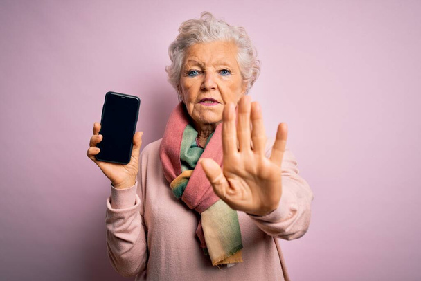 Senior belle femme aux cheveux gris tenant smartphone montrant écran sur fond rose avec main ouverte faisant stop signe avec expression sérieuse et confiante, geste de défense
 - Photo, image
