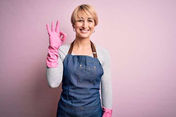 Jeune femme de ménage blonde aux cheveux courts portant tablier et gants sur fond rose souriant positif faisant signe ok avec la main et les doigts. Expression réussie
. - Photo, image