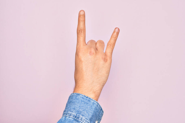 Рука кавказского юноши показывает пальцы на изолированном розовом фоне жестом рок-н-ролла символ, показывающий непристойные рога жест - Фото, изображение