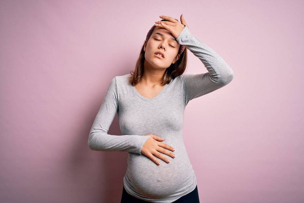 jong mooi tiener meisje zwanger verwachten baby over geïsoleerde roze achtergrond aanraken voorhoofd voor ziekte en koorts, griep en kou, virus ziek - Foto, afbeelding