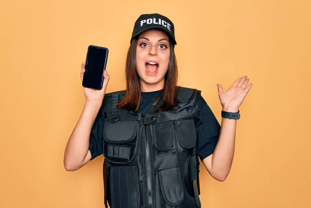 Hermosa mujer policía morena vistiendo uniforme policial sosteniendo el teléfono inteligente mostrando la pantalla muy feliz y emocionada, la expresión ganadora celebrando la victoria gritando con una gran sonrisa y levantando las manos
 - Foto, imagen