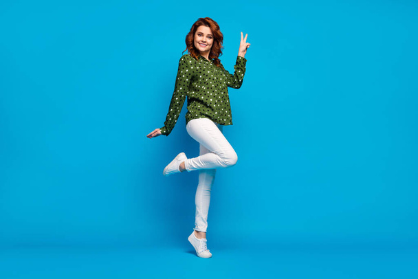 Hi guys. Full length profile photo of pretty cheerful lady good mood flirty person show v-sign symbol wear green dotted shirt white pants footwear isolated blue color background - Foto, Imagem