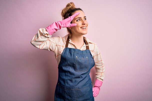 Young beautiful blonde cleaner woman doing housework wearing arpon and gloves very happy and smiling looking far away with hand over head. Searching concept. - Фото, зображення