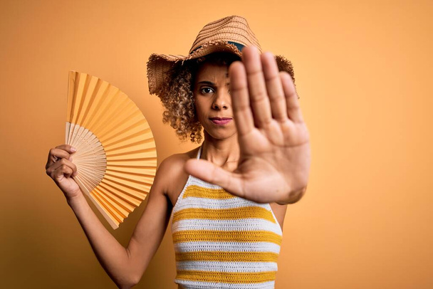 African american tourist woman with curly on vacation wearing summer hat using hand fan with open hand doing stop sign with serious and confident expression, defense gesture - Foto, imagen
