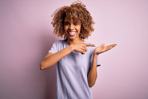 Молодая красивая африканская американка в обычной футболке, стоящей на розовом фоне, поражена и улыбается в камеру, показывая рукой и указывая пальцем.
. - Фото, изображение