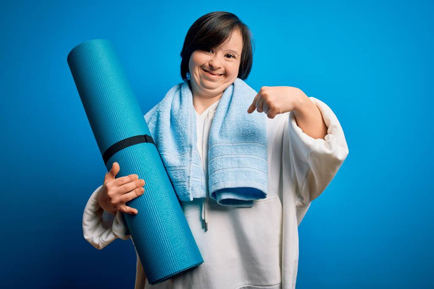 Giovane giù sindrome fitness donna formazione yoga e pilates tenendo tappeto allenamento con faccia a sorpresa puntando il dito verso se stesso
 - Foto, immagini