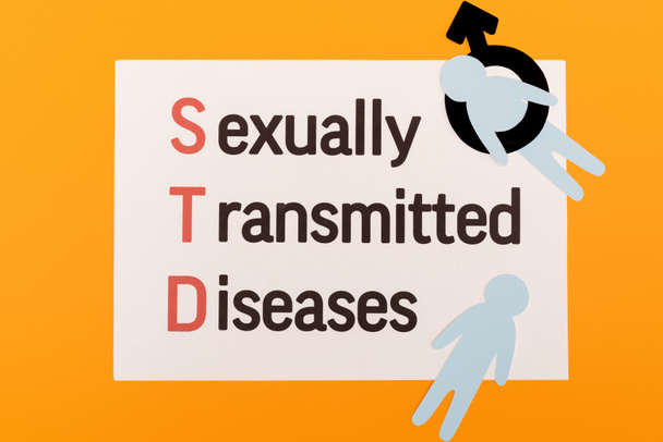 Papiermenschen mit männlichem Geschlechtssymbol in der Nähe von Papier mit sexuell übertragbaren Krankheiten Schriftzug isoliert auf orange - Foto, Bild
