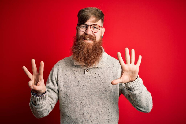Hezký irský ryšavý muž s vousy na sobě ležérní svetr a brýle nad červeným pozadím ukazuje a ukazuje s prsty číslo osm a zároveň se usmívá sebevědomě a šťastně. - Fotografie, Obrázek