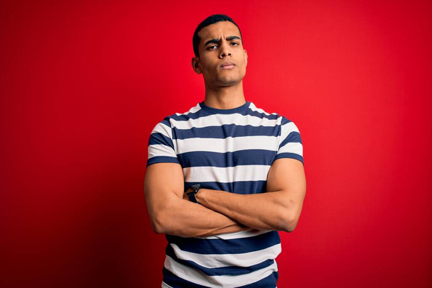 Нахабний афро-американський чоловік у смугастій футболці стоїть над червоним фоном скептично і нервово, не схвалюючи вираз обличчя перехрещеними руками. Негативна людина. - Фото, зображення
