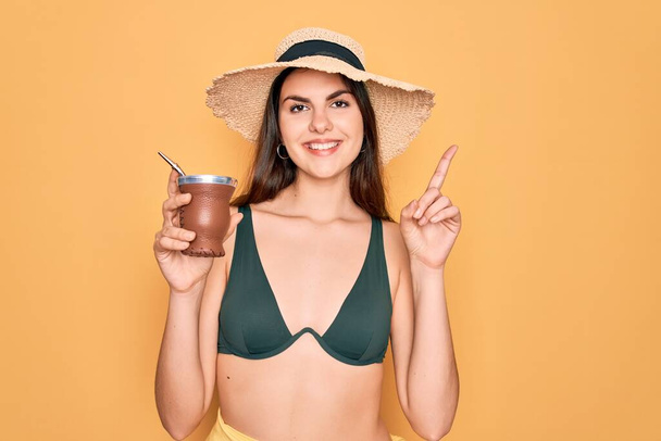 Junge schöne Mädchen in Badebekleidung Bikini und Sommerhut trinken traditionellen Mate-Drink überrascht mit einer Idee oder Frage Zeigefinger mit glücklichem Gesicht, Nummer eins - Foto, Bild