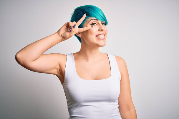 Молодая красивая женщина с голубыми волосами моды в повседневной футболке на белом фоне делает символ мира с пальцами на лице, улыбаясь весело показывая победу
 - Фото, изображение