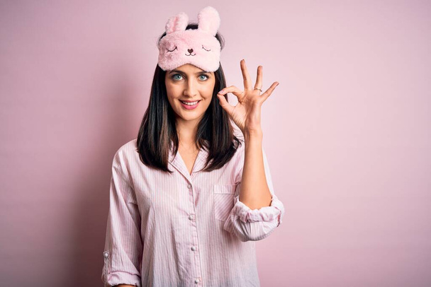 Young brunette woman with blue eyes wearing pink pajama and eye mask over pink background doing ok sign with fingers, excellent symbol - Photo, Image