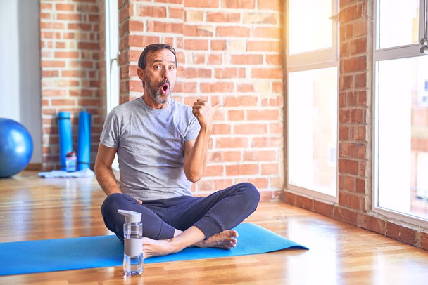 Moyen âge beau sportif assis sur le tapis faire des exercices de yoga étirement à la salle de gym Surpris pointant avec le doigt de la main sur le côté, bouche ouverte expression étonnante
. - Photo, image
