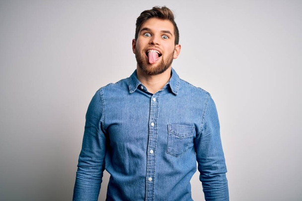 Young handsome blond man with beard and blue eyes wearing casual denim shirt sticking tongue out happy with funny expression. Emotion concept. - Fotoğraf, Görsel