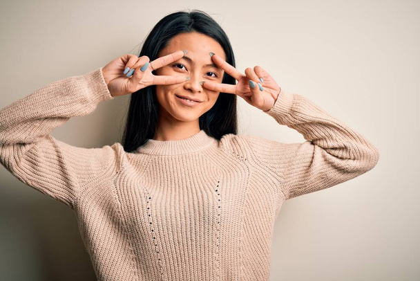 Young beautiful chinese woman wearing casual sweater over isolated white background Doing peace symbol with fingers over face, smiling cheerful showing victory - Foto, imagen