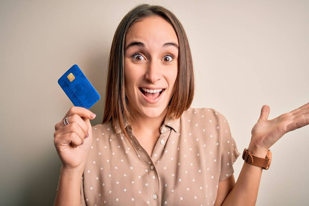 Young beautiful customer woman holding credit card to do payment over white background very happy and excited, winner expression celebrating victory screaming with big smile and raised hands - Photo, Image
