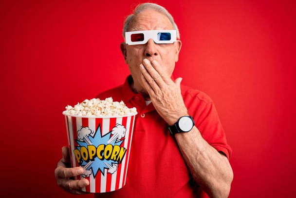 Седовласый пожилой мужчина в 3D очках и ест попкорн над красным фоном, прикрывая рот рукой, потрясенной стыдом за ошибку, выражением страха, испуганной в тишине, секретной концепцией
 - Фото, изображение
