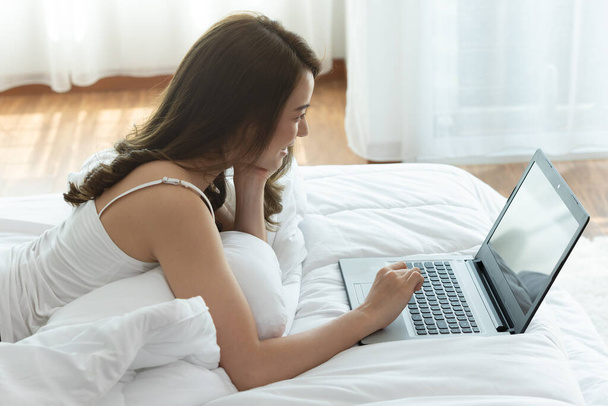 Mujer asiática trabajando en una computadora portátil sentada en la cama en la casa.Mujer revisando aplicaciones sociales y trabajando.Trabajo desde el concepto de casa
. - Foto, Imagen