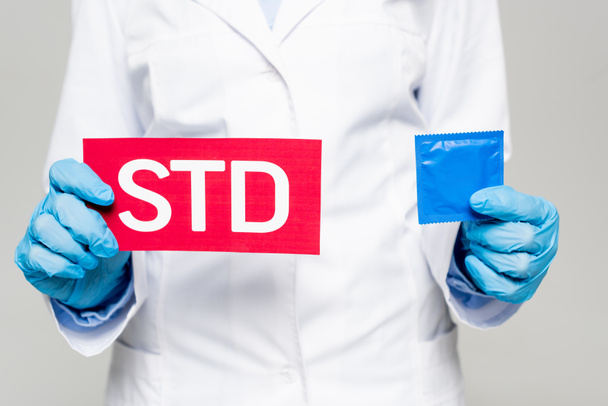 bijgesneden weergave van arts in witte jas en latex handschoenen met papier met STD belettering en condoom geïsoleerd op grijs  - Foto, afbeelding