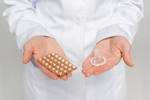 a szürke bőrön izolált fogamzásgátló tablettákat és óvszert tartalmazó buborékcsomagolást tartó orvos levágott képe  - Fotó, kép