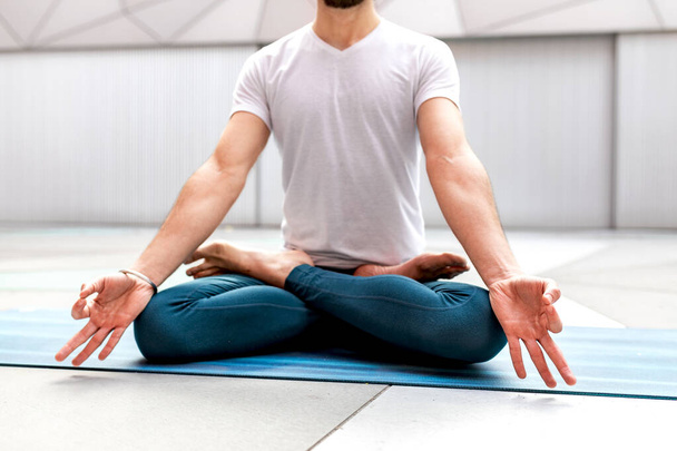 Spor kıyafetleri içinde tanınmayan bir adam Lotus pozisyonunda oturuyor ve kapalı gözlerle meditasyon yapıyor ve geometrik duvarı ve zemini olan geniş bir odada Mudra hareketi yapıyor. - Fotoğraf, Görsel