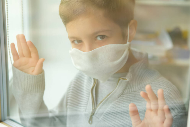 Young boy in a medical mask looks out the window. Self-isolation in quarantine, coronavirus, covid 19. - Fotoğraf, Görsel