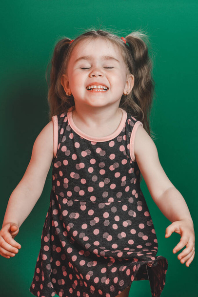 Κοριτσάκι τριών ετών με ουρά σε φόρεμα ποζάρουν στο στούντιο σε πράσινο φόντο 2021 - Φωτογραφία, εικόνα