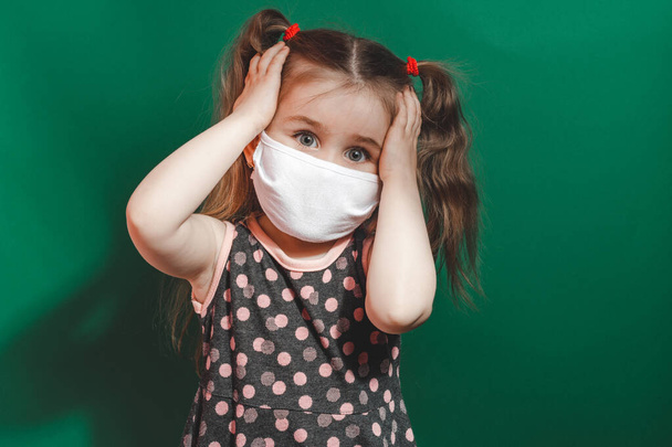 Μικρό καυκάσιο κορίτσι με ιατρική μάσκα φοράει κόκκινο πουά φόρεμα στο στούντιο σε πράσινο φόντο και κρατά το κεφάλι της στον πόνο 2021 - Φωτογραφία, εικόνα