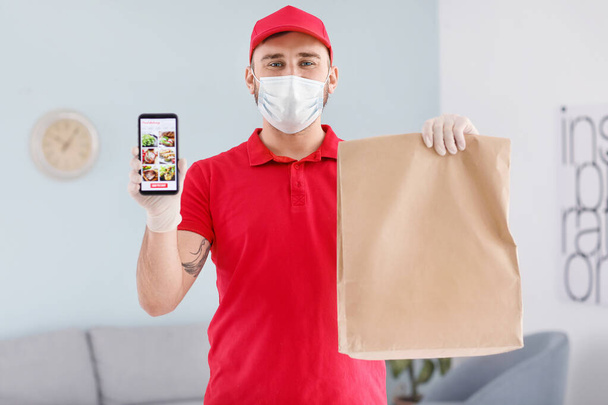 Male courier of food delivery company in medical mask and with mobile phone indoors - Foto, afbeelding