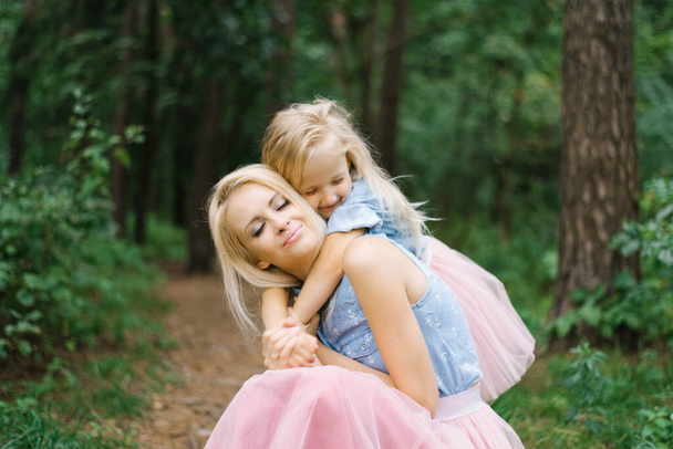 Äiti ja viisivuotias tytär identtisissä vaaleanpunaisissa tyllihameissa ja sinisissä farkkupaidoissa kävelevät puistossa tai metsässä. Tytär halaa äitiään. Äitienpäivä - Valokuva, kuva