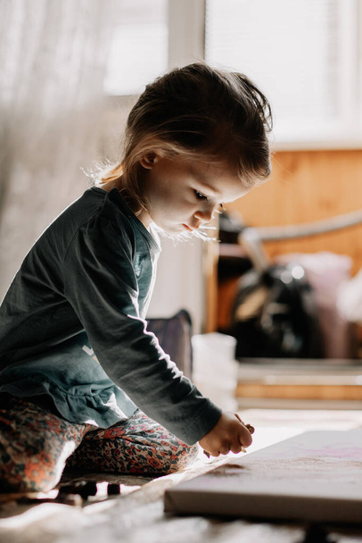 25.06.2018 Vinnitsa, Ukrane : une petite fille dessine des peintures à l'huile sur toile avec des os et un couteau à palette assis sur le sol près de la fenêtre
 - Photo, image