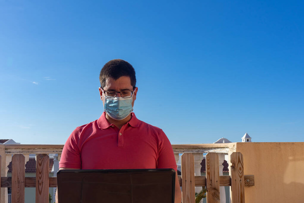 Man met een masker werkend met zijn laptop vanaf het balkon van zijn huis. Hij zit in een leunstoel. Hij draagt een zalmkleurig poloshirt en bril.. - Foto, afbeelding