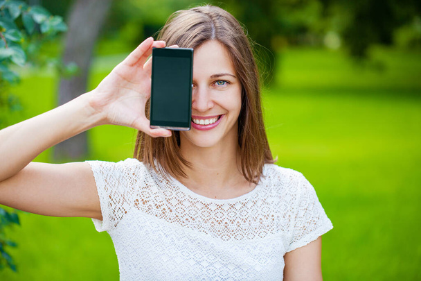 Young beautiful brunette woman showing your smartphone screen, summer park outdoors - Fotoğraf, Görsel