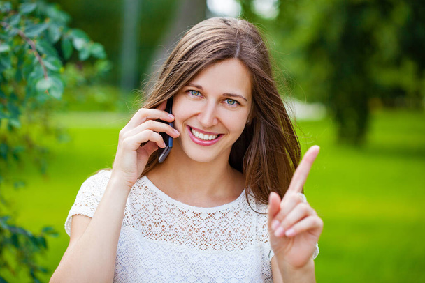 Portrait of beautiful brunette young woman speaking on mobile phone at summer green park - Zdjęcie, obraz