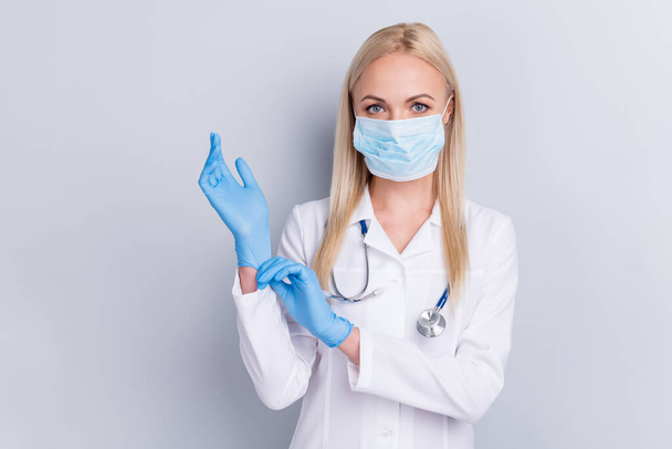 Close-up portrait of nice attractive blonde girl paramedic surgeon medic expert specialist assistant phonendoscope stethoscope putting rubber gloves on isolated on grey pastel color background - 写真・画像
