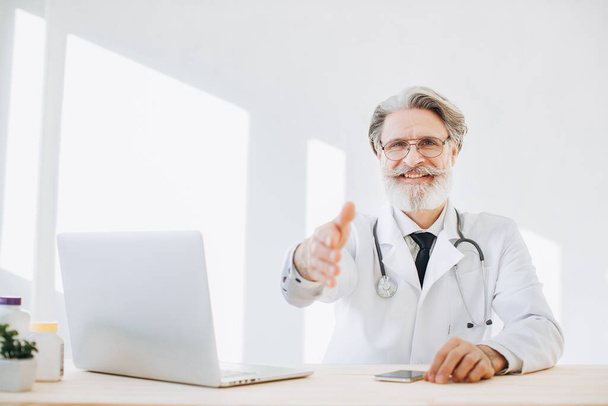 Bearded senior physician sitting at a desk and offering handshake - Фото, зображення