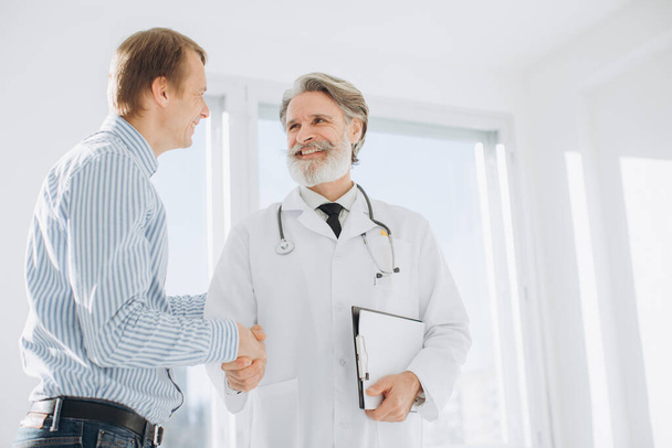 Médico mayor estrechando la mano con el paciente mientras se reúne en el consultorio del médico
. - Foto, imagen