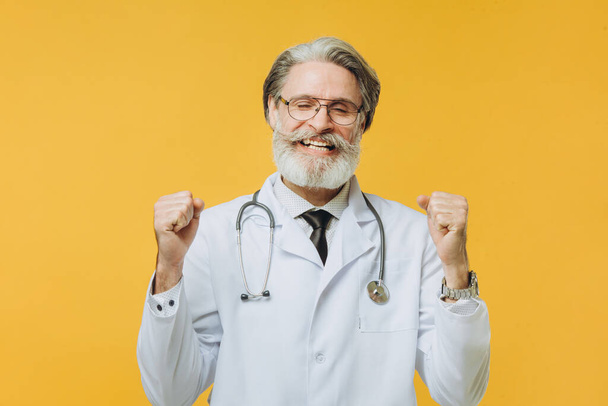 Senior grey-haired doctor man wearing stethoscope standing over isolated yellow background celebrating happy and amazed for success. Winner concept. - Fotografie, Obrázek