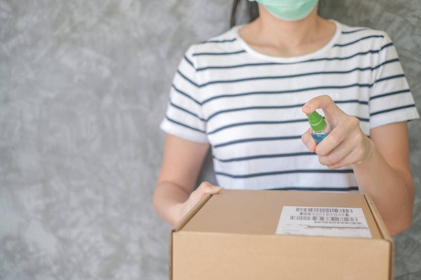 Frau mit Schutzmaske mit Sprühalkoholdesinfektionsmittel reinigt Paketpost, um Viren und Bakterien vom Postabsender zu verhindern schützt sie sich und ihre Familie vor Virus Frau gibt Paket - Foto, Bild