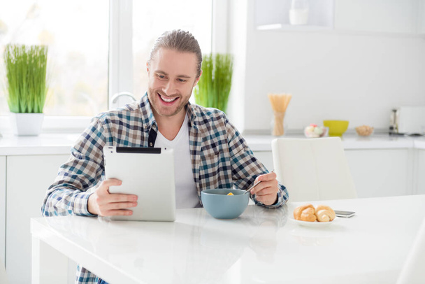 Portrait of his he nice attractive cheerful cheery glad focused guy holding in hand tablet watching video film blog vlog eating snack lunch alone in modern light white interior style kitchen - Fotó, kép
