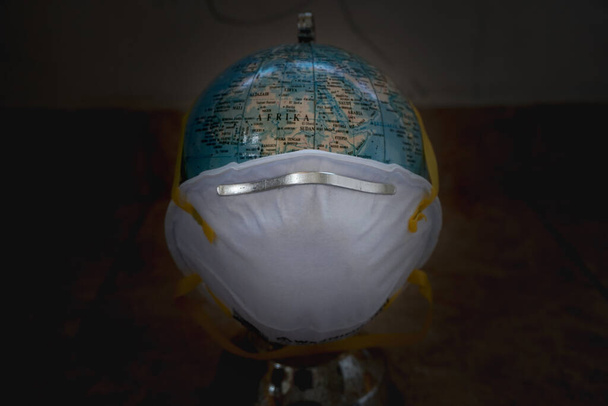 un globo terrestre simulado cubierto con una máscara N95, lo que significa que el mundo está siendo atacado por la pandemia covid-19. Fondo oscuro ayuda a centrarse en objetos globo y máscaras n95 - Foto, Imagen