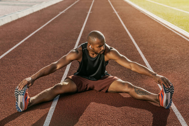 Sportowy muskularny afrykański sportowiec w słuchawkach rozciągające nogi podczas siedzenia na torze wyścigowym stadionu - Zdjęcie, obraz
